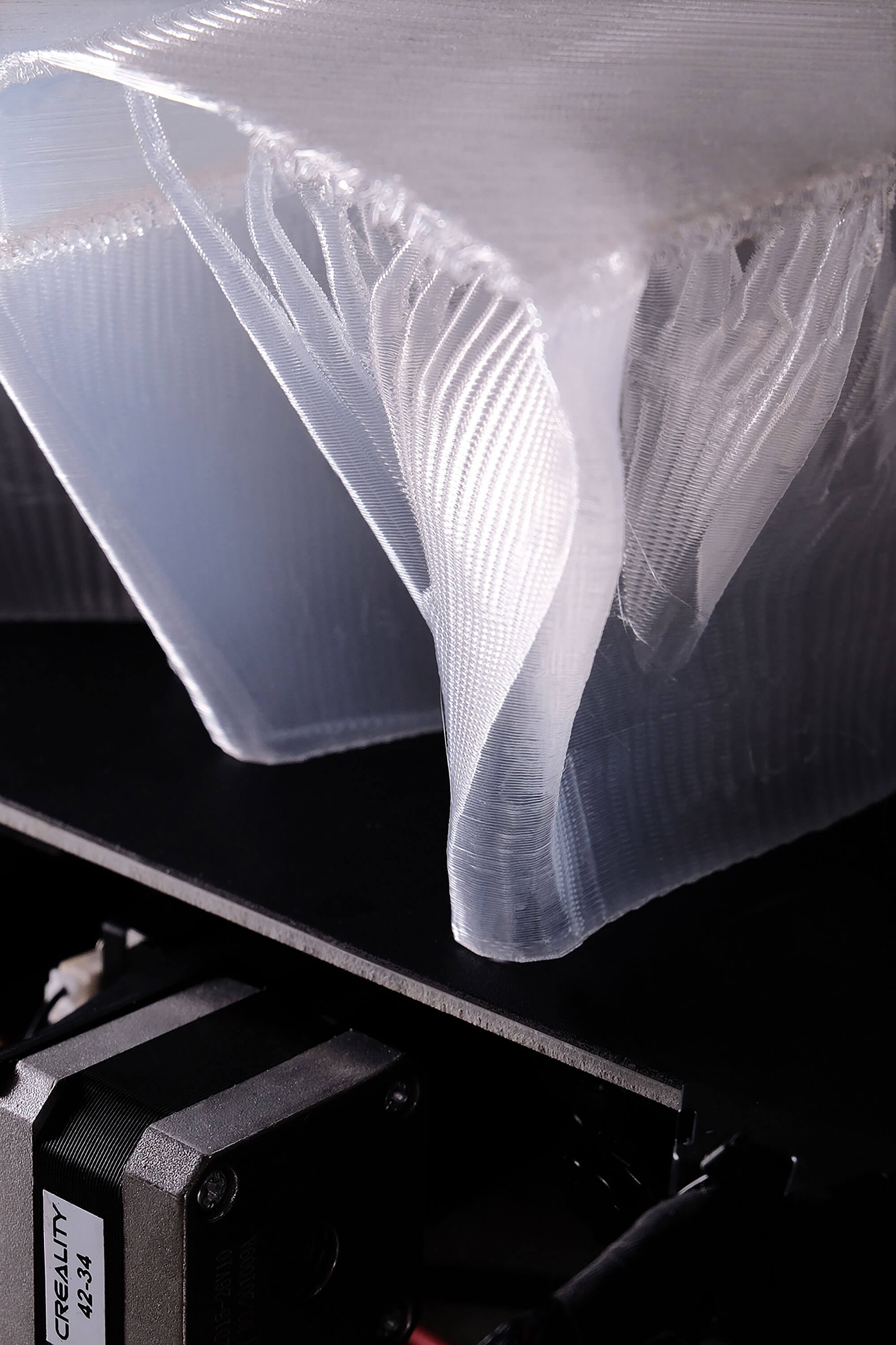 Close-up of a translucent, 3D printed book with layered, curved ridges, resting on a black platform. Below it, part of a CREALITY 3D printer and a stepper motor are visible.